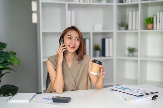A young professional smiles during a phone call in her well-organized office, holding a coffee cup, reflecting a blend of personal engagement and business communication.