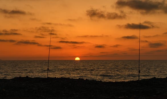 Two fishing rods stand tall against the vibrant sunset, as waves gently lap at the rocky shoreline, creating a peaceful atmosphere perfect for evening leisure.