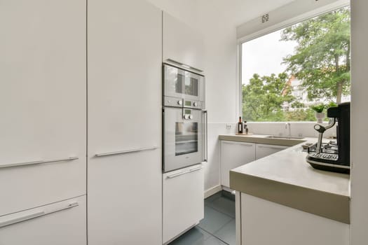 Bright and spacious kitchen featuring sleek, white cabinetry, modern appliances, and large windows revealing greenery outside, creating a serene cooking environment.