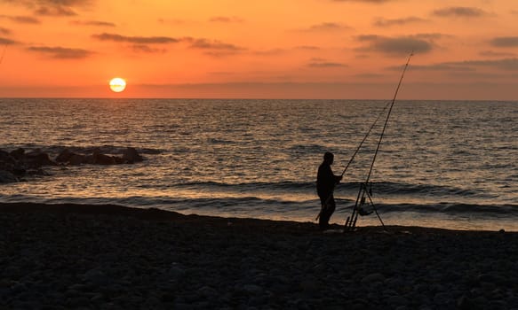A silhouette of a dedicated fisherman stands on a rocky shore as the sun sets, painting the sky with warm hues. The rhythmic sound of waves completes this peaceful moment.