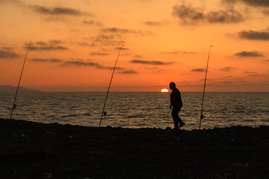 A lone figure walks along the rocky shoreline, silhouetted against the vibrant hues of a setting sun. Fishing poles rise from the ground, capturing the serenity of the evening.