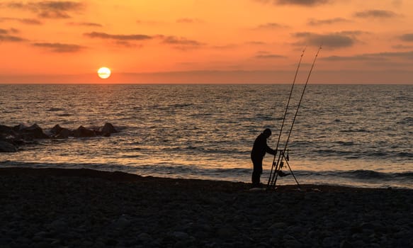 A lone fisherman stands on a pebbled beach, silhouette outlined against a breathtaking sunset. Rods in hand, he embraces the calm of the evening, surrounded by tranquil waters.