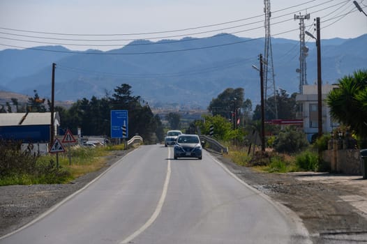 Vehicles cruise along a winding road surrounded by lush greenery and rolling hills, with majestic mountains in the backdrop under a bright blue sky. Peaceful countryside vibes abound.
