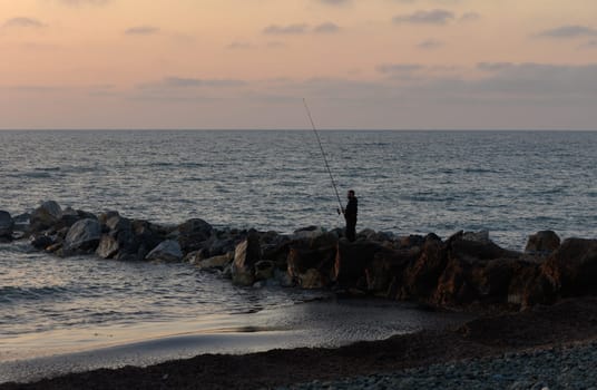 Silhouetted against a pastel sky, a lone fisherman stands on rocky shores, casting his line into the calm waters during the tranquil dusk hour.