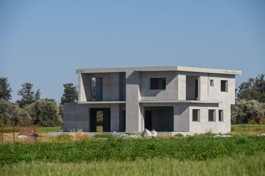 Amidst lush green fields, a minimalist concrete house stands tall, showcasing its modern architecture. The structure reflects contemporary design principles on a sunny day.