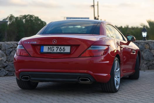 Gaziveren Cyprus -16.04.2025 A stunning red sports car stands gracefully against a picturesque sunset backdrop. The sun casts a soft glow, showcasing the car's glossy finish and elegant lines as it relaxes by a tranquil setting.