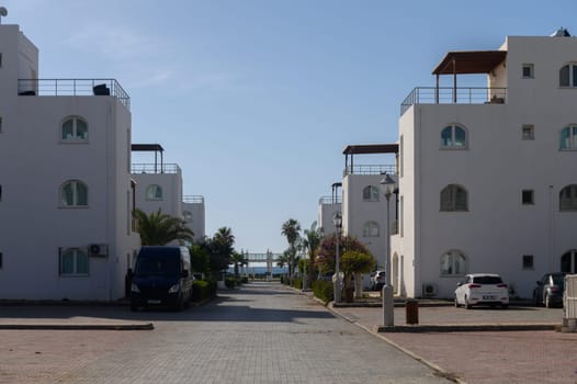 Whitewashed buildings line a peaceful avenue, leading to the shimmering sea on a bright day. Lush palms sway gently, adding to the serene coastal atmosphere.