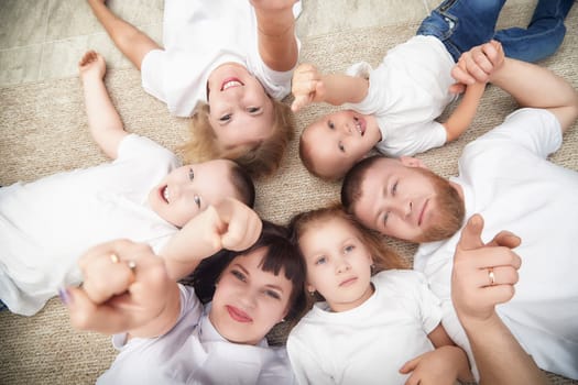 Six family members are lying on a rug, smiling and pointing at the camera, enjoying a fun moment together in a warm indoor setting.