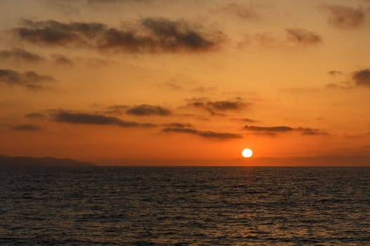 As the day transitions to night, warm golden and orange tones paint the sky above the ocean. Gentle waves ripple as the sun slowly sinks below the horizon, creating a serene atmosphere.