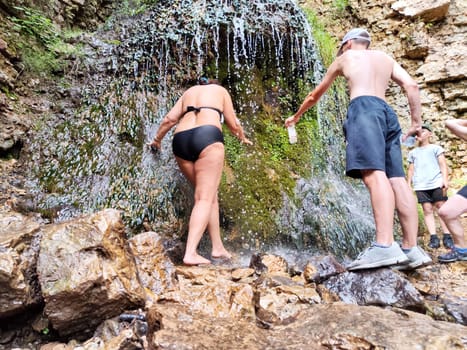 Friends enjoy a cooling experience under a waterfall in a rocky setting during a summer hike, surrounded by nature's beauty.