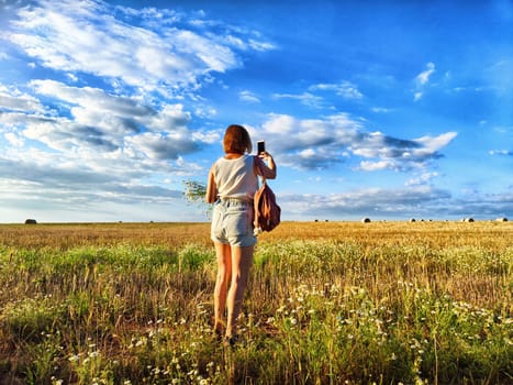 A young woman stands in a sunlit field holding flowers and capturing a selfie against a backdrop of blue skies and clouds.