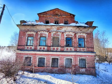 An abandoned brick structure sits under a clear blue sky, surrounded by snow in a desolate rural area during the winter season.