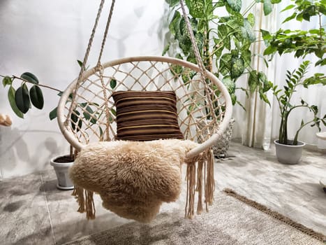 A hammock chair hangs in a bright, green indoor space, adorned with a textured pillow and soft throw, surrounded by lush plants.