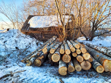 Stacked wooden logs lie next to a rustic cabin covered in snow, surrounded by a serene winter landscape and bare trees.