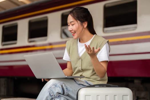 A young woman smiles while using her laptop to connect with friends, blending travel with technology at a train station.