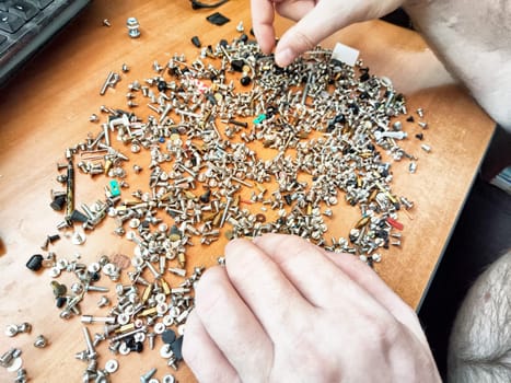 Hands are meticulously sorting a vast collection of metal screws and fasteners on a wooden table in a workshop setting.
