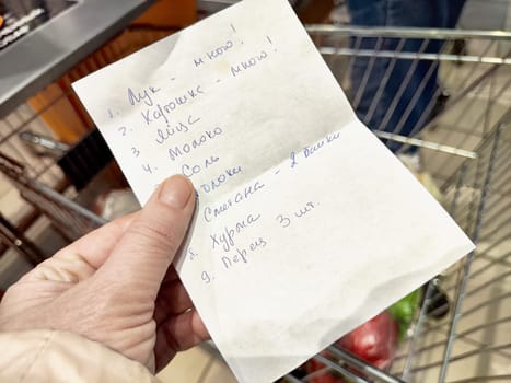 A shopping list in a hand is shown, featuring several food items needed for cooking, held in a grocery cart.