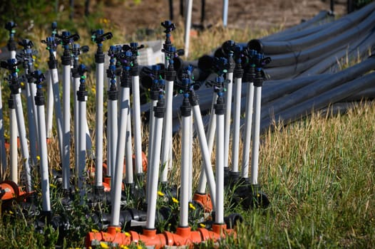 Sprinklers stand ready in an open field, surrounded by vibrant green grass, while irrigation hoses lie coiled nearby, under the warm summer sunlight, preparing for the upcoming watering task.