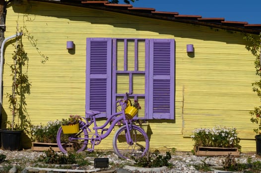 A charming purple bicycle, filled with blooming flowers, leans against a bright yellow wall with matching shutters under a clear blue sky. This playful decor brightens the garden scene.