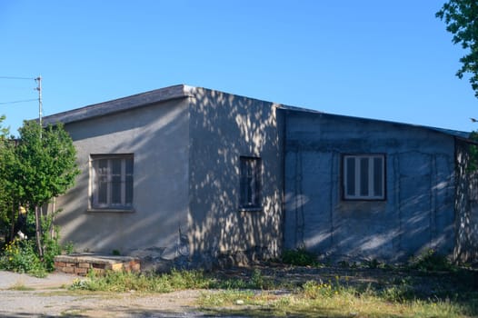 In a serene countryside setting, a weathered building stands under the clear blue sky. Overgrown grass and delicate flowers surround its textured walls, suggesting stories of the past.