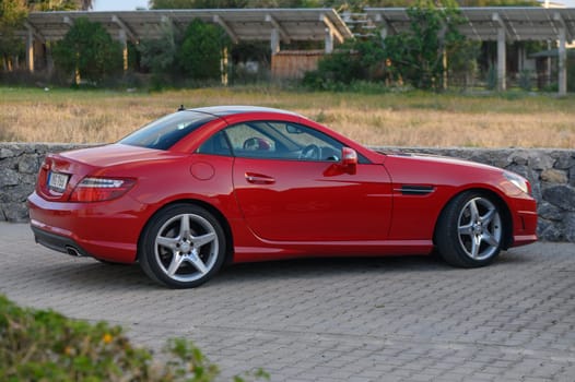 Gaziveren Cyprus -16.04.2025 A striking red sports car is elegantly parked on a paved surface, surrounded by lush greenery under clear blue skies. The sun casts a warm glow, enhancing this serene ambiance.
