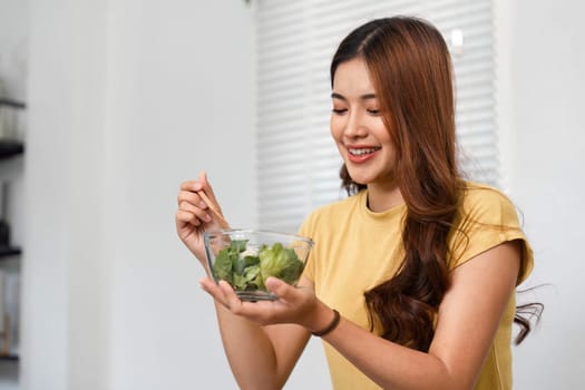 A young woman is seated at home, happily savoring a bowl of salad. Her morning routine includes a nutritious meal, reflecting a commitment to health and wellness.
