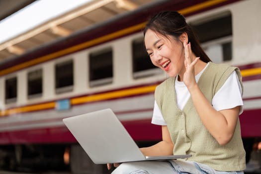 A joyful traveler sits at the train station, engaging in a video call on her laptop, illustrating the blend of travel and remote work in modern life.