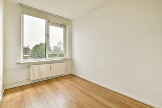 A spacious minimalist room featuring a large window offering a view of a cityscape and greenery outside. The wooden floor enhances the simplicity of the interior.