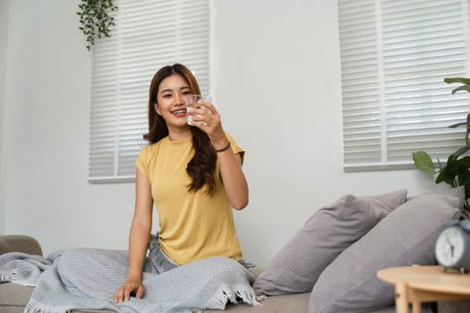 A cheerful young woman sits on the couch, holding a glass of water, ready to indulge in a refreshing morning ritual, emphasizing hydration.