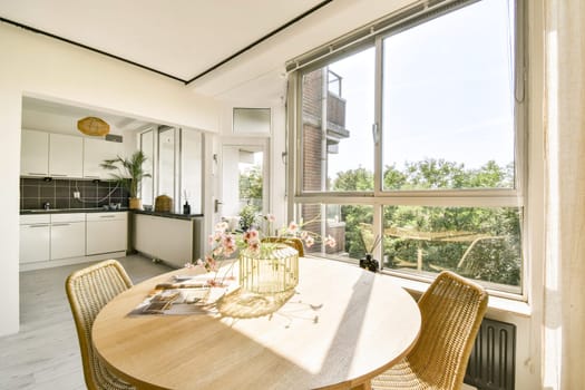 A sunlit dining area featuring a wooden table surrounded by woven chairs, with large windows offering views of greenery, perfect for a cozy dining experience.