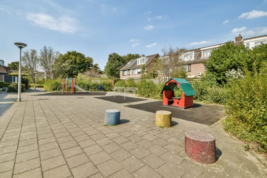 A colorful playground featuring a slide and a playhouse, surrounded by greenery and modern residential buildings under a clear blue sky.