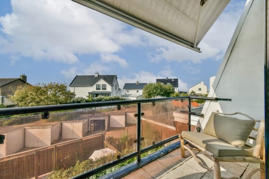 A serene balcony scene featuring a stylish chair and a view of residential houses surrounded by lush greenery and blue skies.