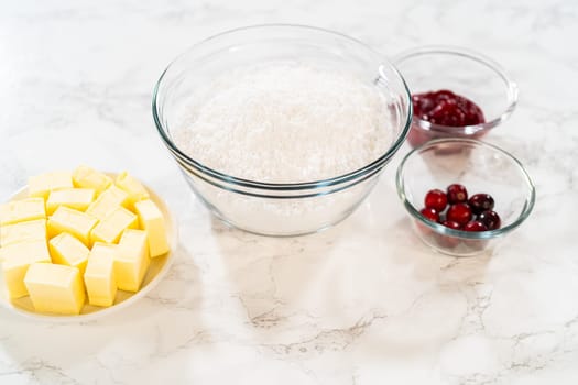 An overhead view of the essential ingredients for a baking recipe, featuring cubed butter, a bowl of all-purpose flour, and fresh cranberries. The butter is neatly arranged in cubes, while the flour is stored in a large glass bowl. The cranberries are shown in two smaller bowls, ready to be incorporated into the recipe. This visually pleasing setup emphasizes fresh, high-quality ingredients, perfect for food blogs, recipe development, or ingredient-focused content.