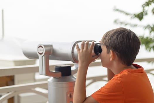 Child in orange shirt using a mounted public telescope on an outdoor observation deck with railing and tree in the background