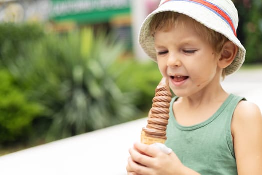 Young boy in green shirt and striped hat holding and licking chocolate ice cream cone on a sunny day