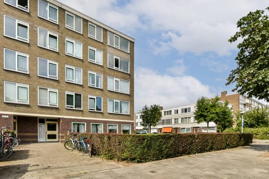 A contemporary building featuring large windows, surrounded by trees and manicured shrubs, with bicycles parked in front, showcasing urban living.