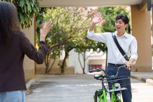 A cheerful young man on a bicycle waves to a friend as they pass each other in a leafy neighborhood, promoting sustainable transportation and friendly connections.