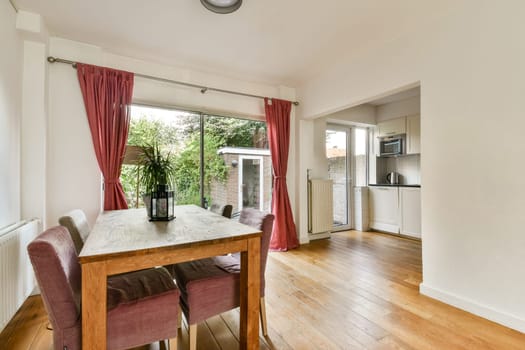 A contemporary dining space featuring a wooden table and chairs, bathed in natural light from large windows, with greenery visible outside.