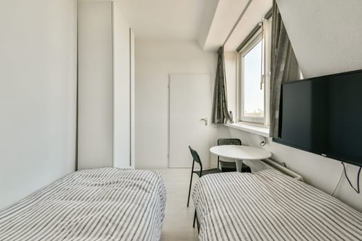 A bright and minimalist bedroom featuring two single beds, a table, and a television with large windows providing ample natural light.