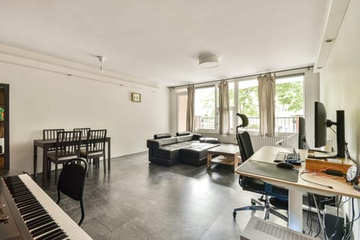 A spacious modern living room featuring a comfortable sofa, dining table, and a dedicated workspace with a computer, large windows allowing natural light.