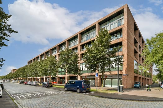A contemporary building features large glass windows surrounded by lush greenery and trees, set on a bustling urban street with parked vehicles.