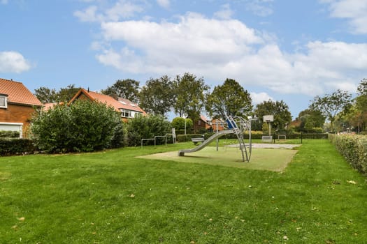 A peaceful park scene showcasing a playground surrounded by lush greenery and residential homes under a clear blue sky.