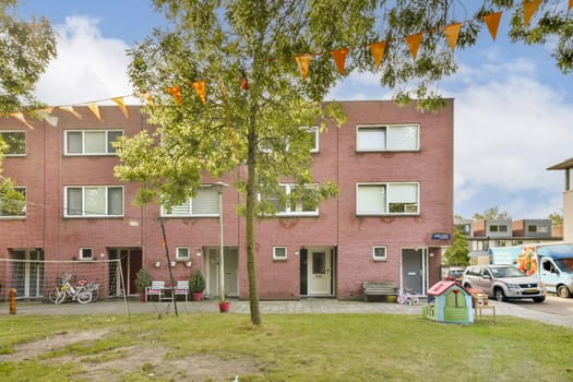 A vibrant residential scene featuring a red brick building, trees, and festive decorations. Bicycles and children’s play equipment add to the inviting atmosphere.