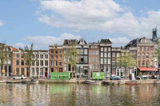 Colorful canal houses line the waterway in Amsterdam, showcasing classic Dutch architecture with reflections shimmering in the water under a blue sky.