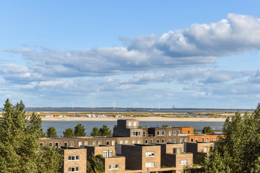 A picturesque landscape showing modern buildings alongside a tranquil waterfront, complemented by lush greenery under a clear blue sky.