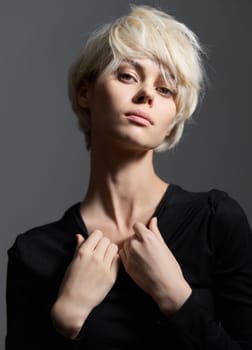 Elegant female model with short platinum blonde hair, wearing a fitted black top. Standing against a dark gray background, she portrays strength and self-assurance through her intense gaze and postur