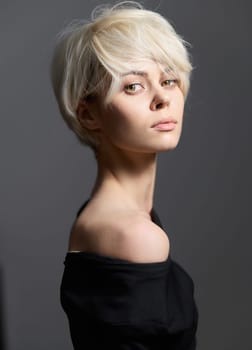 Confident model with short platinum blond hair gazing into the camera, wearing a chic off-shoulder black top against a soft gray background. Art and fashion theme.