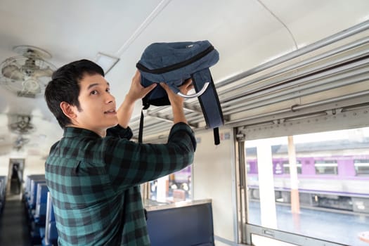 A train passenger organizes his belongings by storing his backpack above him, ready for the adventure ahead. His excitement is palpable.