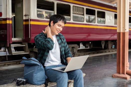 While waiting for his train, a young traveler enjoys the comfort of technology, working on his laptop with a smile.