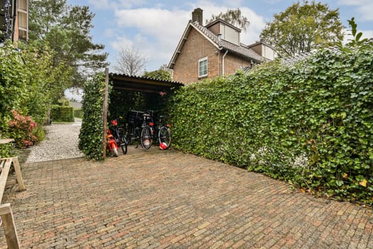 A picturesque garden pathway lined with greenery and brick paths, featuring parked bicycles under a shaded area. Nature and architecture blend harmoniously.